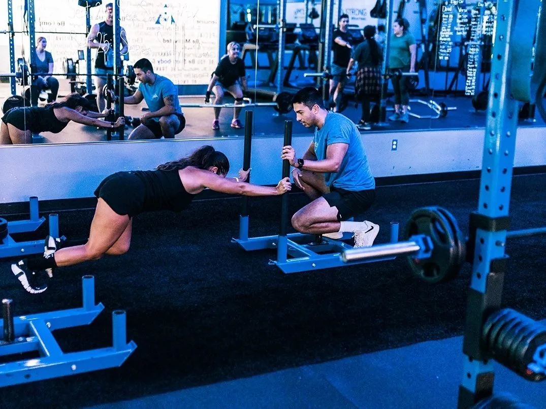 Coach Santi kneeling beside a client, coaching her one-on-one through a sled push and adjusting the plan to her body in real time